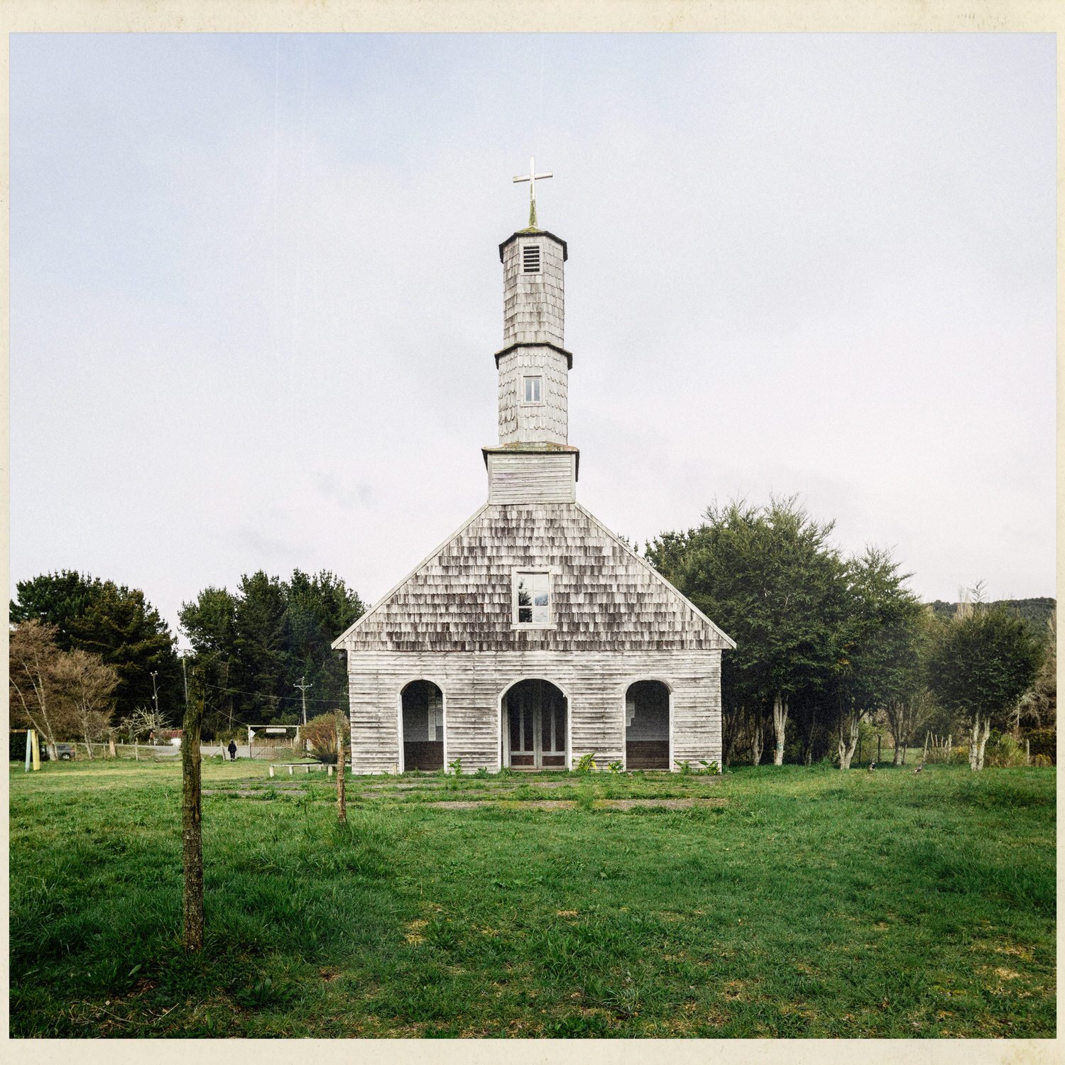 Fachada de la iglesia de Compu con su torre alta y tres arcos, frente a un prado