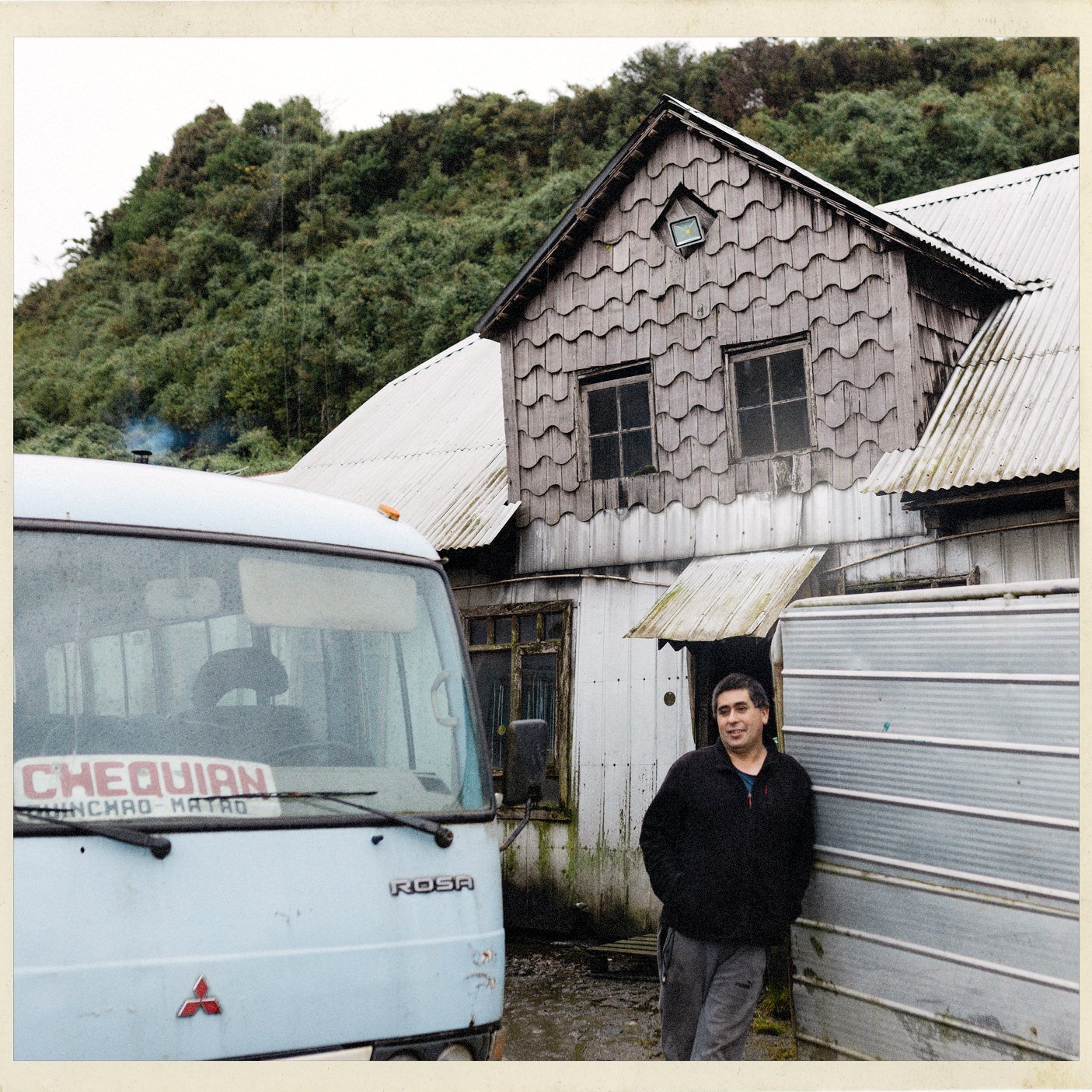 Wladimir de pie junto a un bus rural y una casa de tejuelas