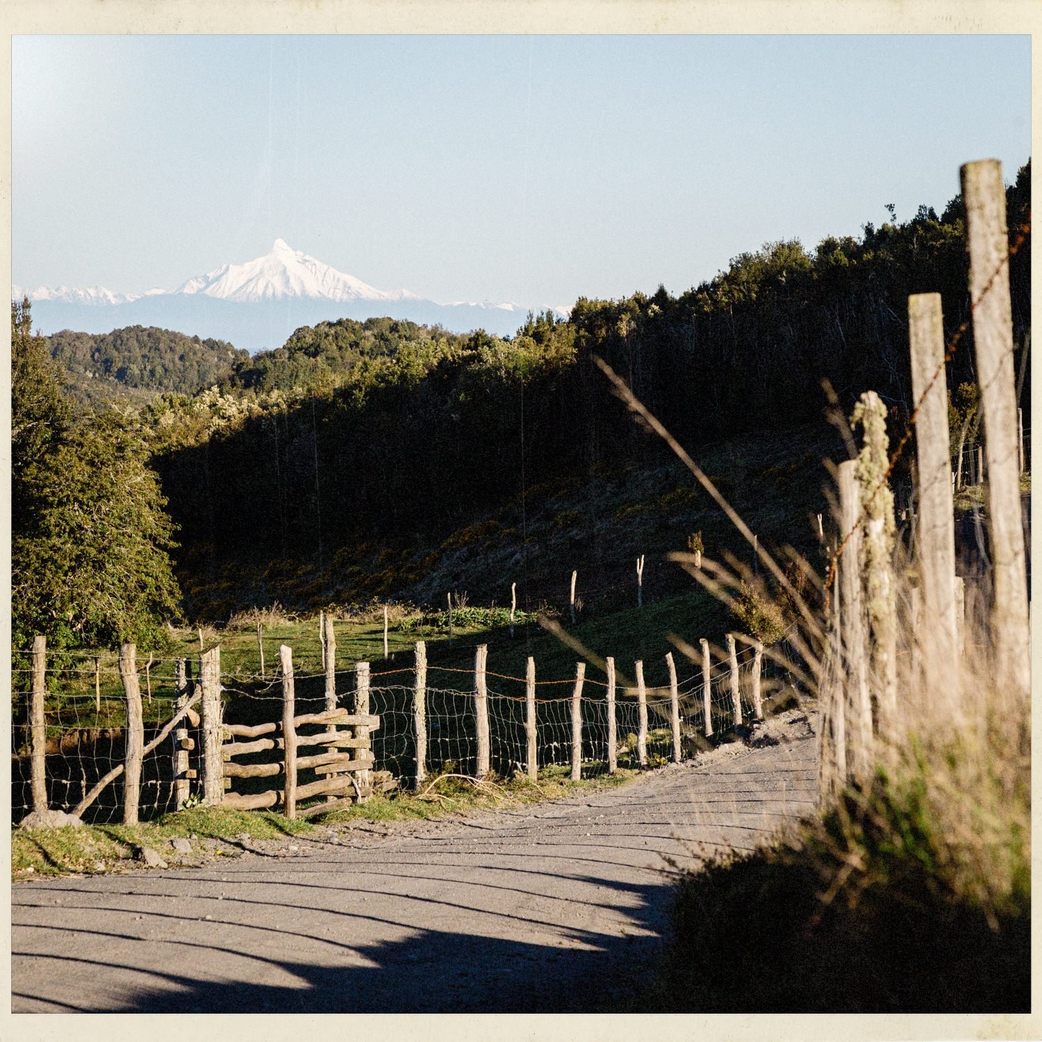 Camino rural con cercos de madera y un volcán nevado al fondo