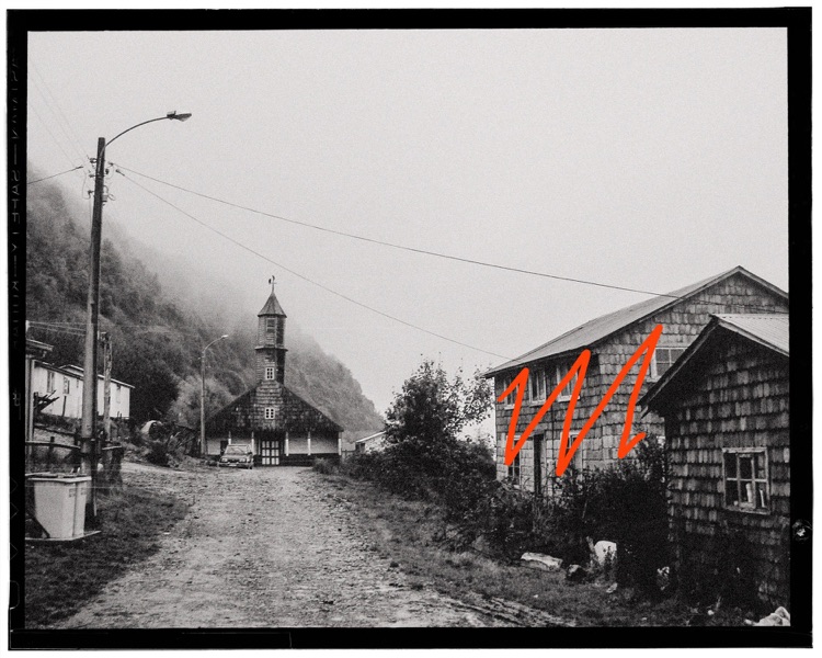 Vista en blanco y negro de una calle rural con casas de madera, un árbol, una iglesia en el fondo y montañas cubiertas de niebla.