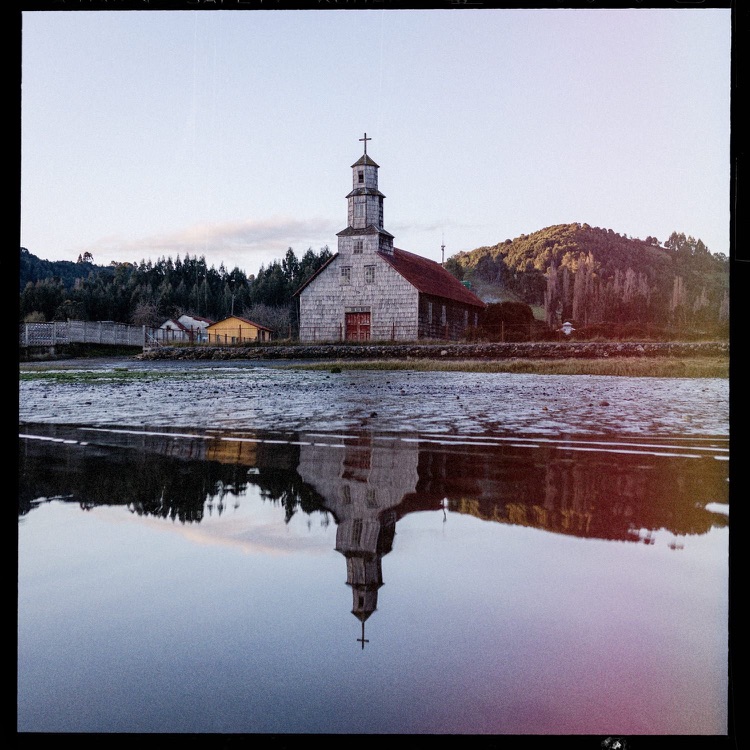 Una iglesia de madera antigua junto a un lago con reflejo de la estructura y un fondo de colinas y árboles