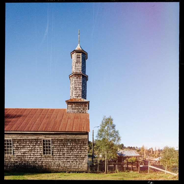 Iglesia de madera con techo de lámina roja y torre del reloj, rodeada de árboles y cielo claro.