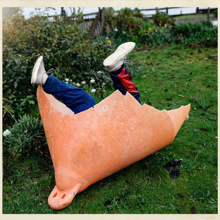 Niño de espaldas con pantalones deportivos y zapatos deportivos, saltando dentro de una cáscara de huevo gigante, en un jardín con césped y arbustos.
