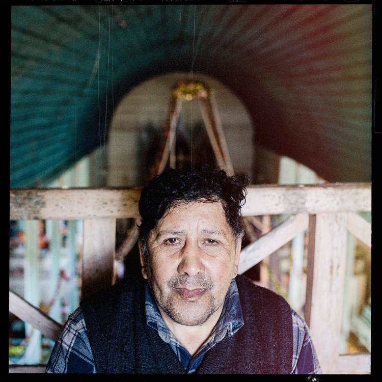 Retrato de un hombre con barba y cabello rizado oscuro, con expresión neutral, sentado frente a una estructura de madera. Fondo con un túnel o pasa…