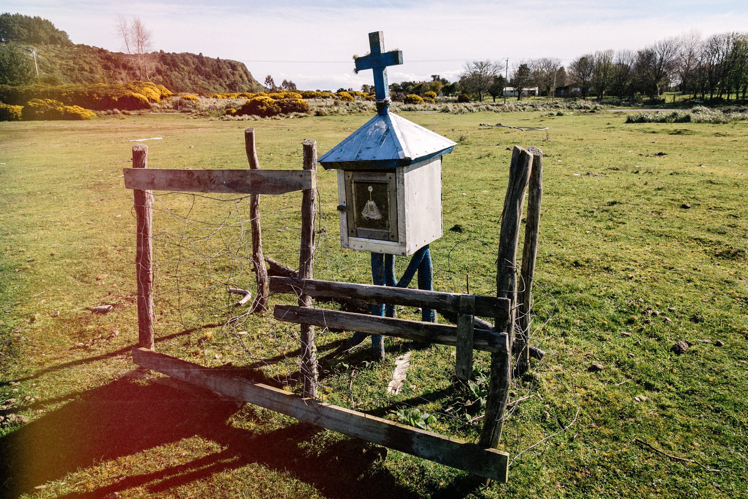 Pequeño santuario de madera con cruz junto a un cerco, en un campo de Apiao