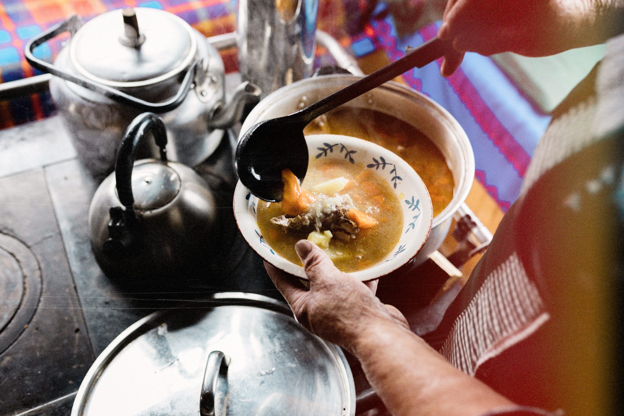 Manos sirviendo cazuela junto a una cocina a leña con teteras