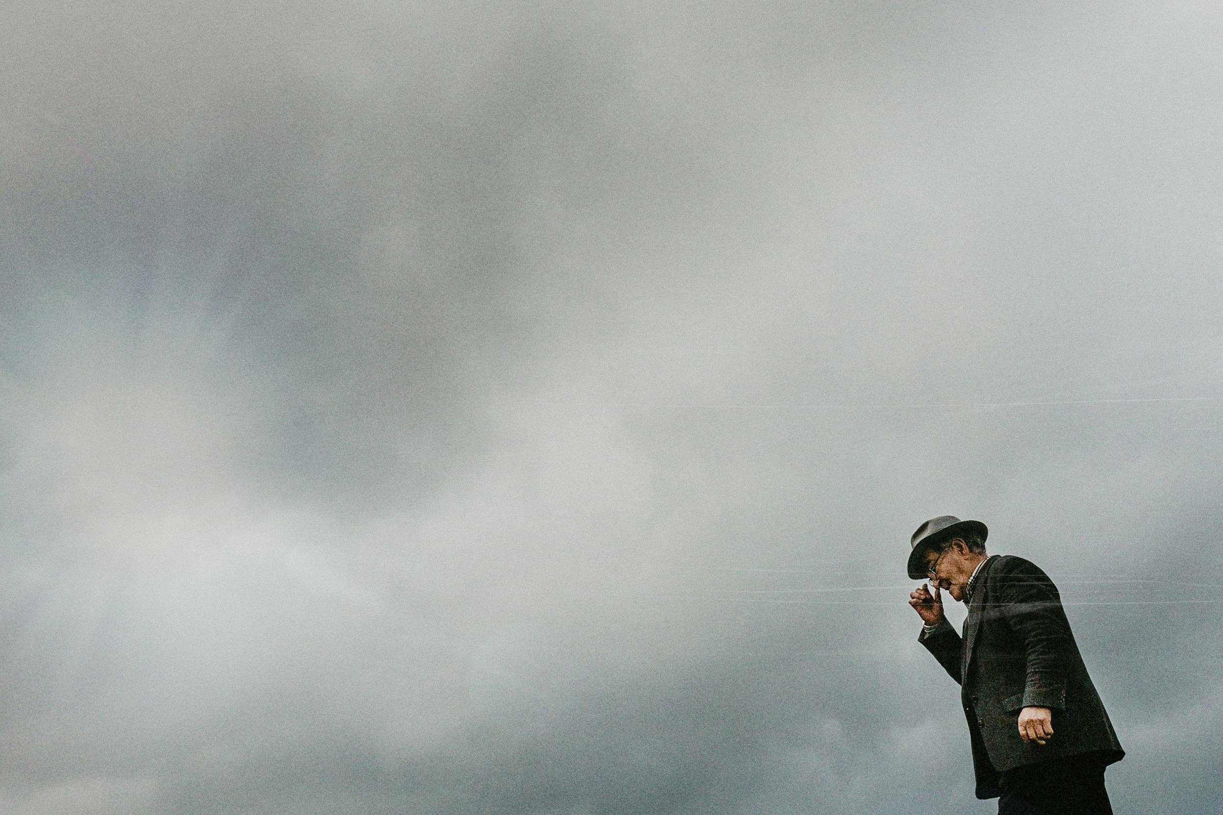 Hombre con sombrero, gafas y traje oscuro mirando hacia abajo y con la mano en la frente, en un cielo nublado y gris.